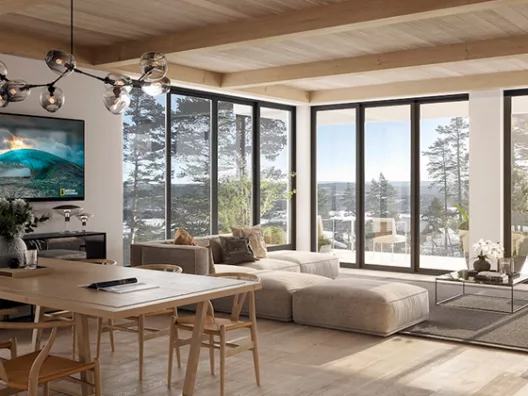 Moderne stue med store vinduer som viser utsikt over skogen, møblert med sofa, spisebord og TV som viser et havbilde.