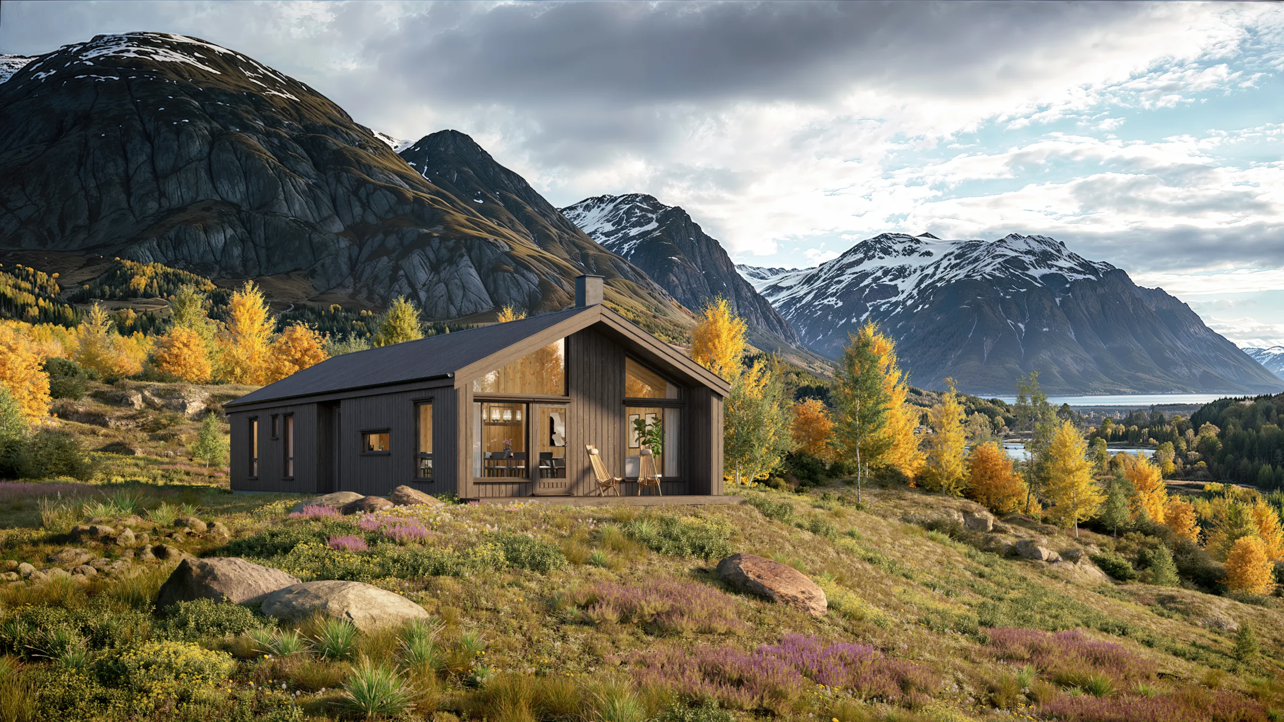 En moderne trehytte ligger i en gressbakke med fjell, høsttrær og en innsjø i bakgrunnen under en delvis overskyet himmel.