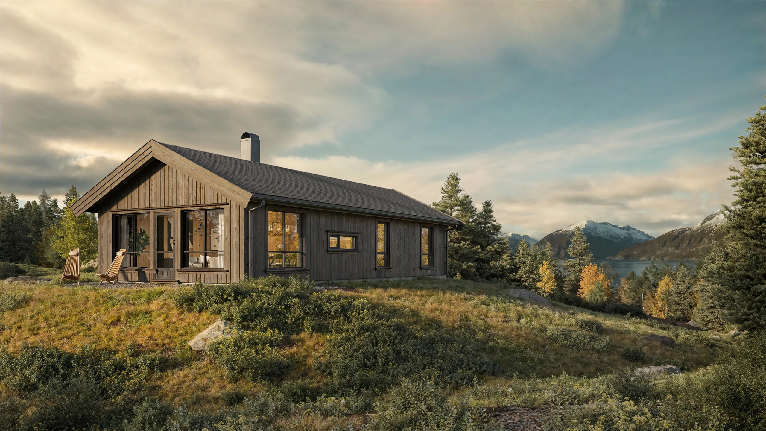 En trehytte med store vinduer ligger på en gressbakke omkranset av trær, med fjell og en innsjø synlig i bakgrunnen under en delvis overskyet himmel.