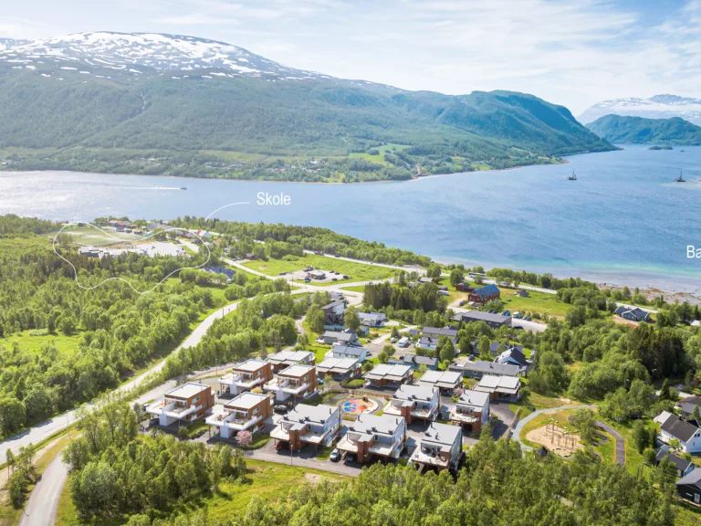 Flyfoto av en liten by nær en fjord med fjell i bakgrunnen; etiketter viser hvor en skole og en barnehage ligger.