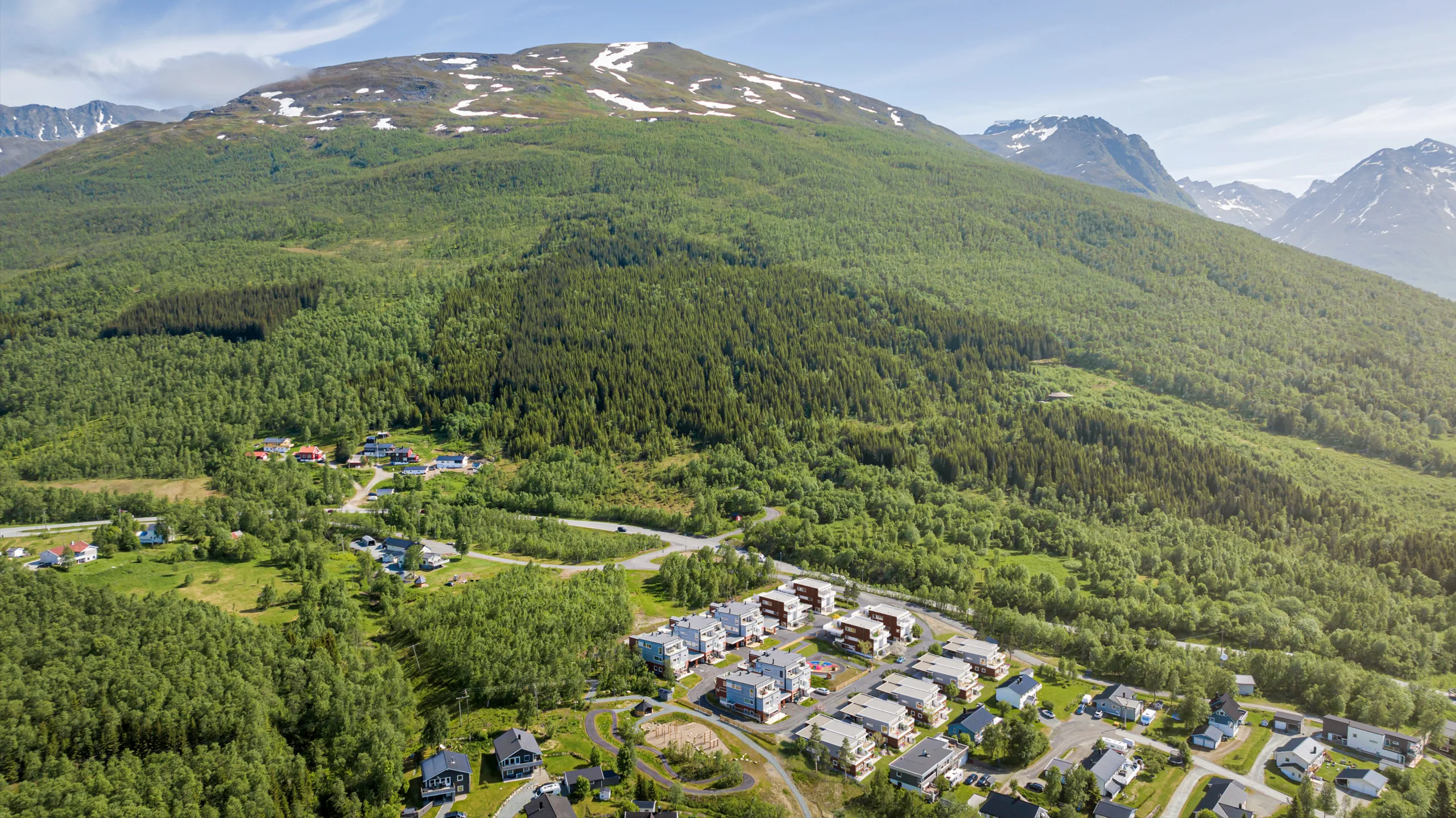 Flyfoto av en liten by med spredte hus ved foten av et skogkledd fjell, med snøflekker nær toppen.
