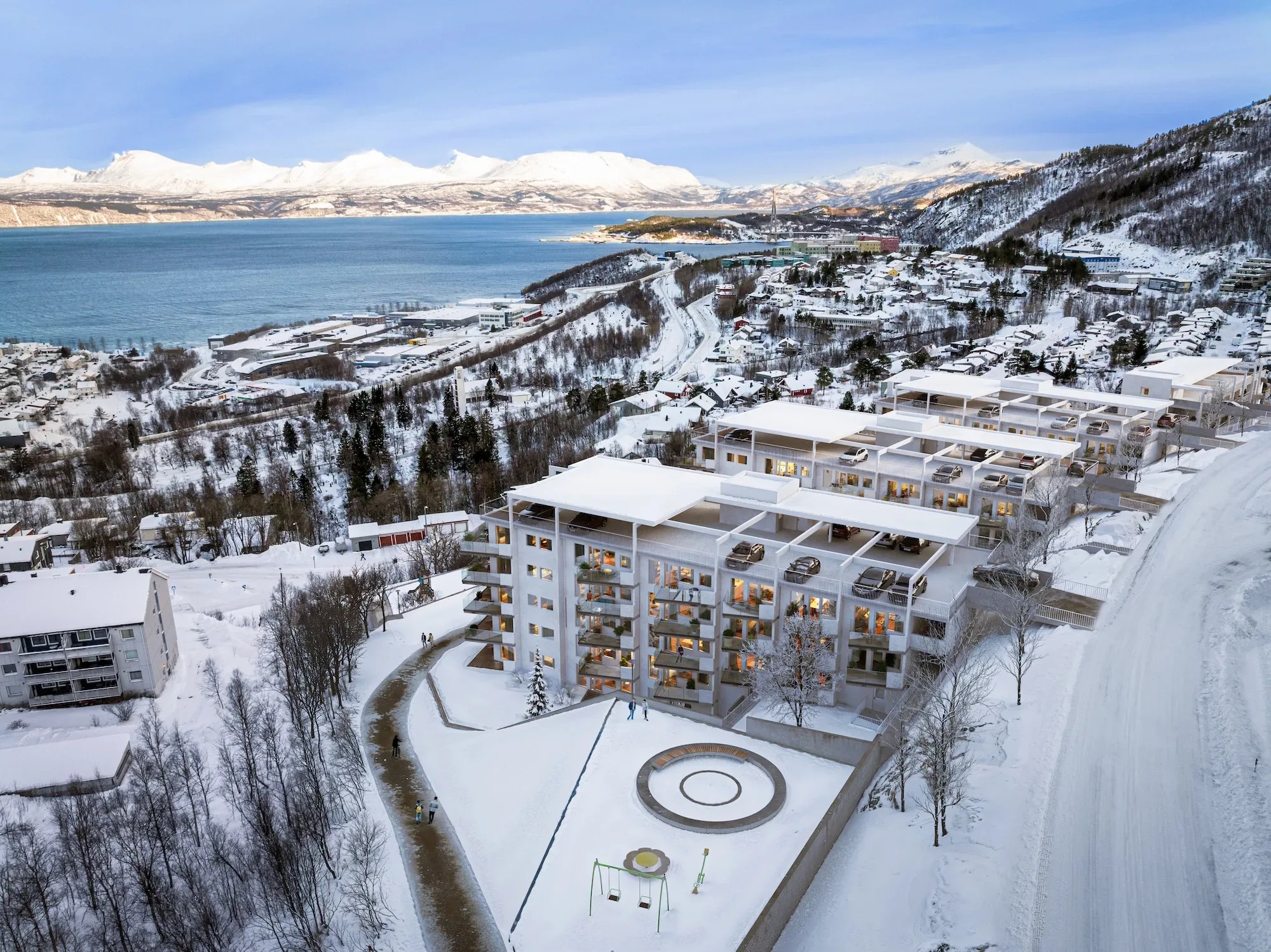 Flyfoto av et moderne leilighetskompleks omgitt av snø med fjell og vann i bakgrunnen.