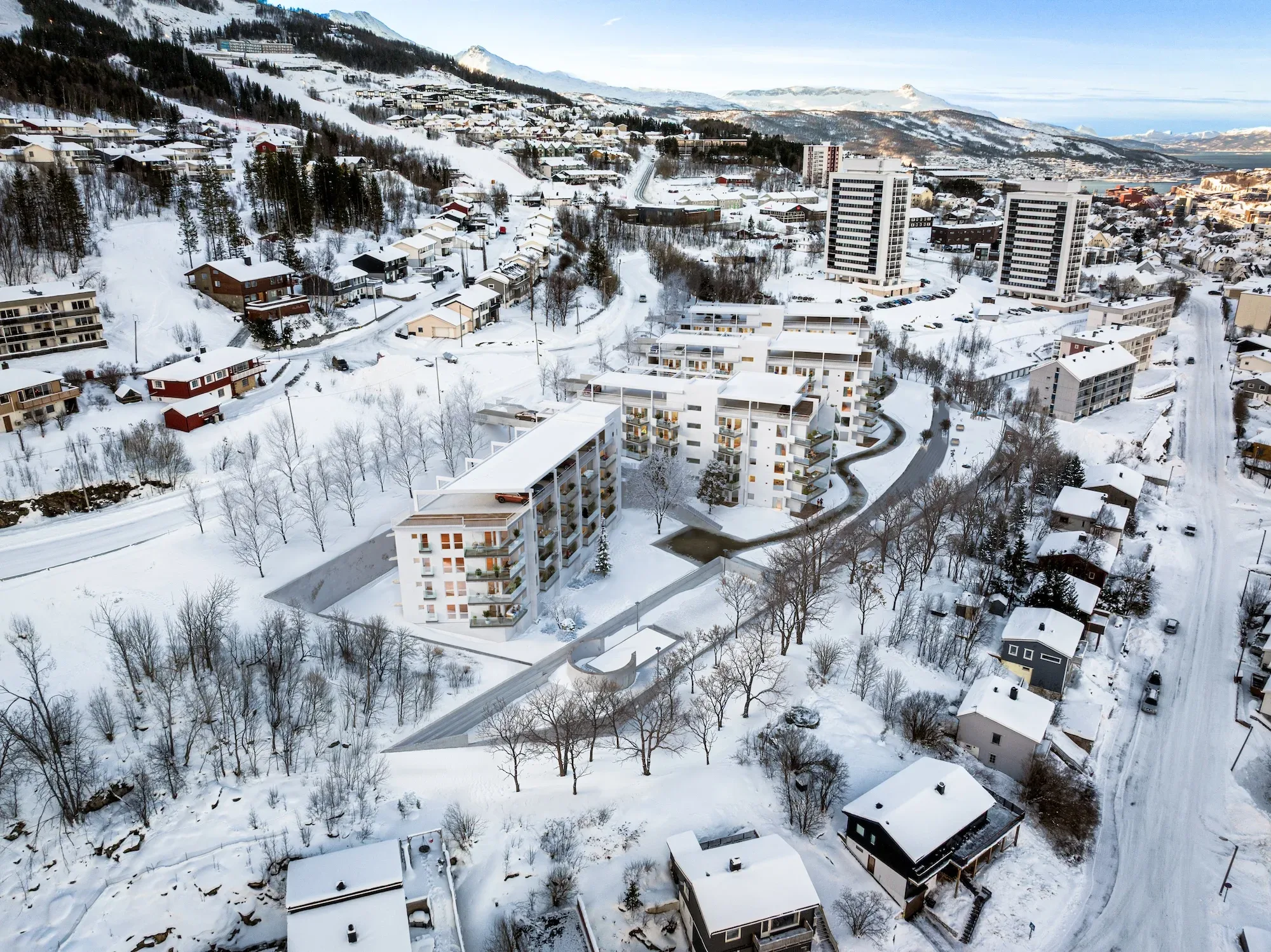 Flyfoto av et snødekt boligområde med moderne bygårder, mindre hus og gater med trær i et kupert landskap.