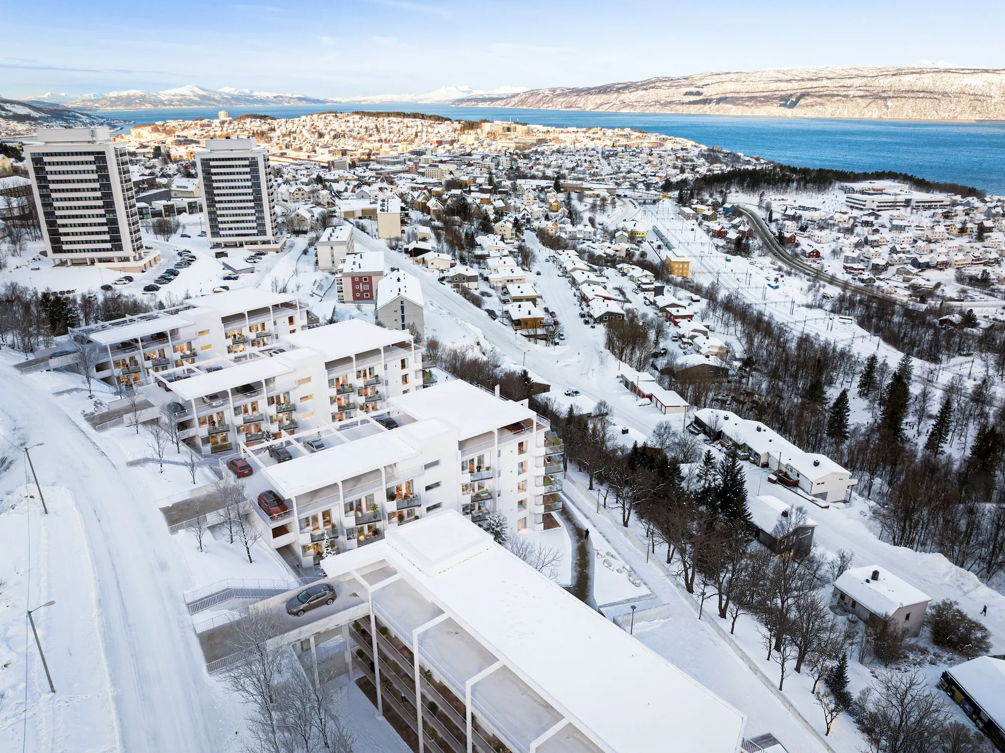 Flyfoto av et snødekt bylandskap med boligblokker, høyhus og et vann i bakgrunnen, omgitt av snødekte trær og veier.