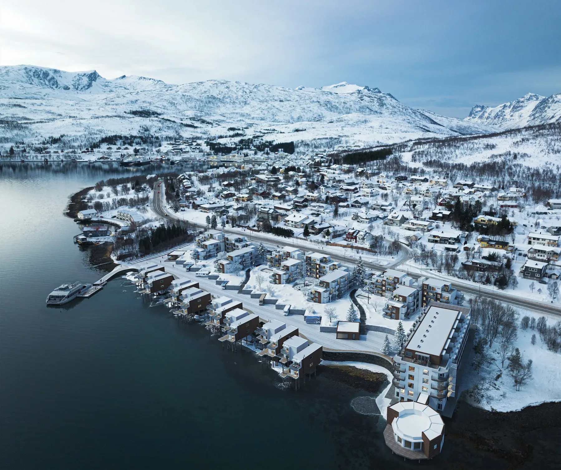 Flyfoto av en snøkledd by ved innsjøen med moderne bygninger langs strandlinjen, omgitt av fjell og delvis frosset vann.