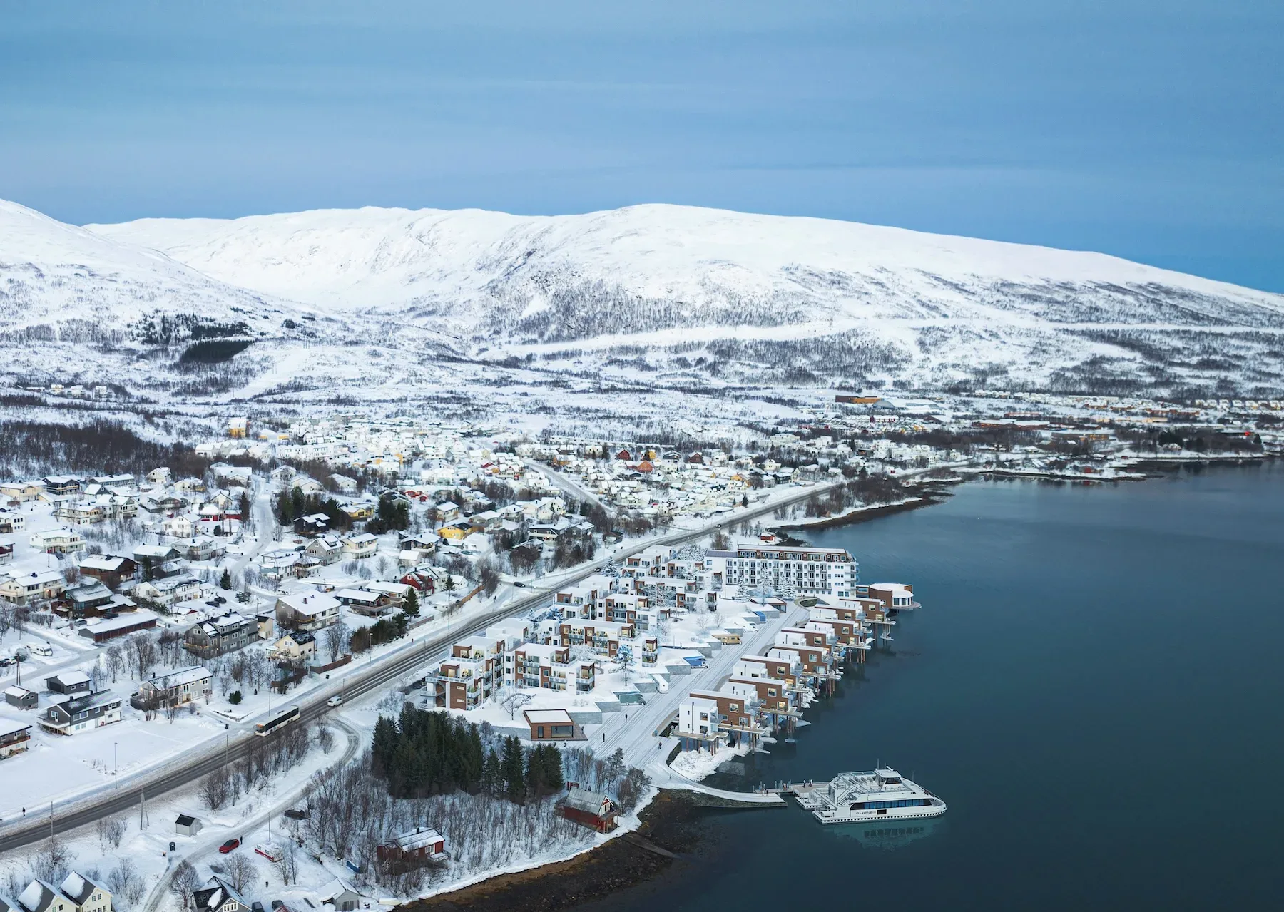 Flyfoto av en snødekt kystby med moderne bygninger ved vannet, et fartøy som ligger til kai, og snødekte fjell i bakgrunnen.