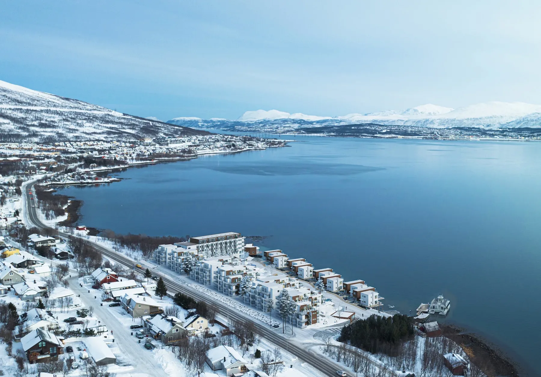 Flyfoto av en snødekt kystby med bygninger langs strandlinjen, et rolig, blått vann og fjell i bakgrunnen under en klar himmel.